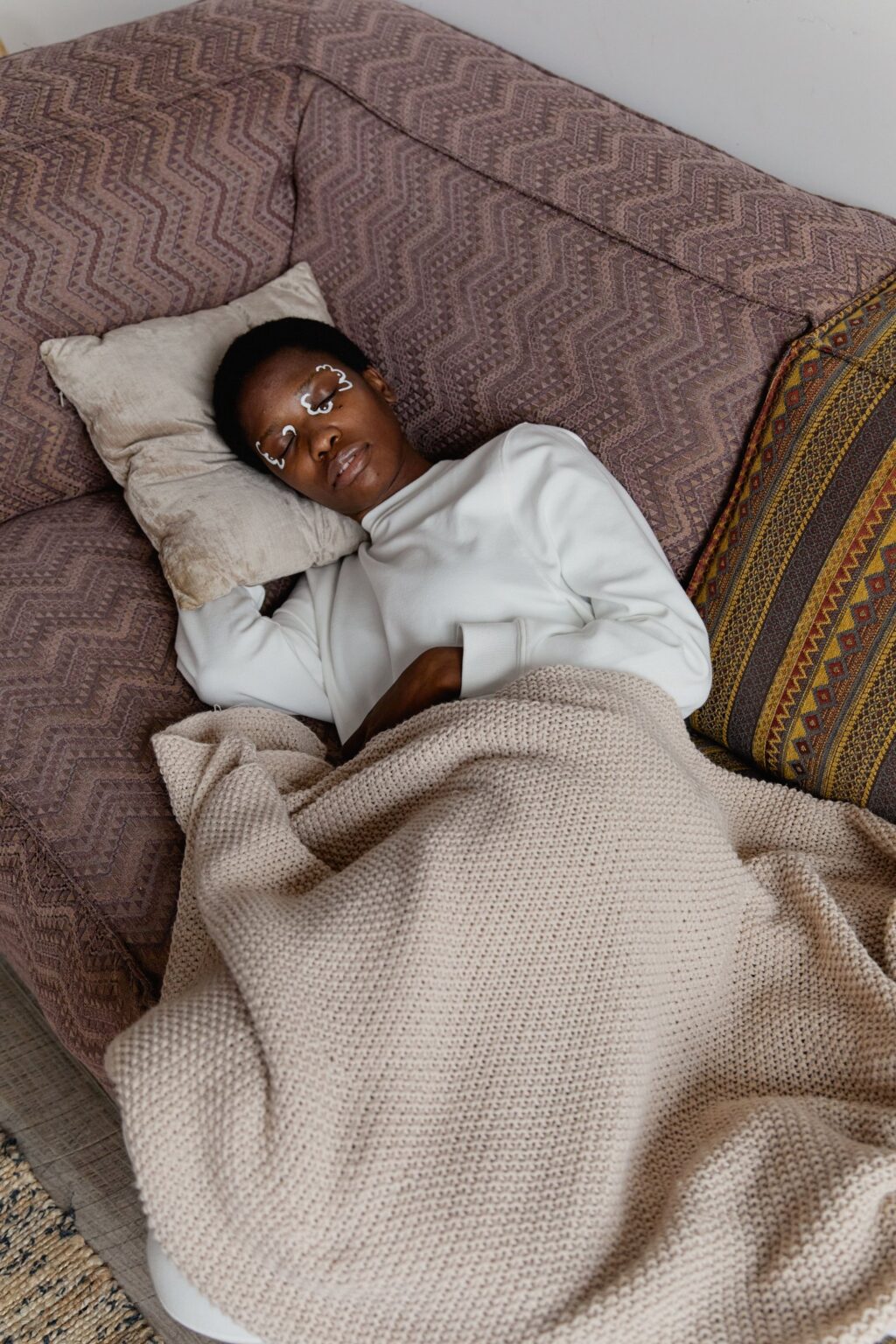 Sleeping teenager rests on a patterned sofa, tucked beneath a beige knitted blanket with a pillow and colorful striped cushion nearby