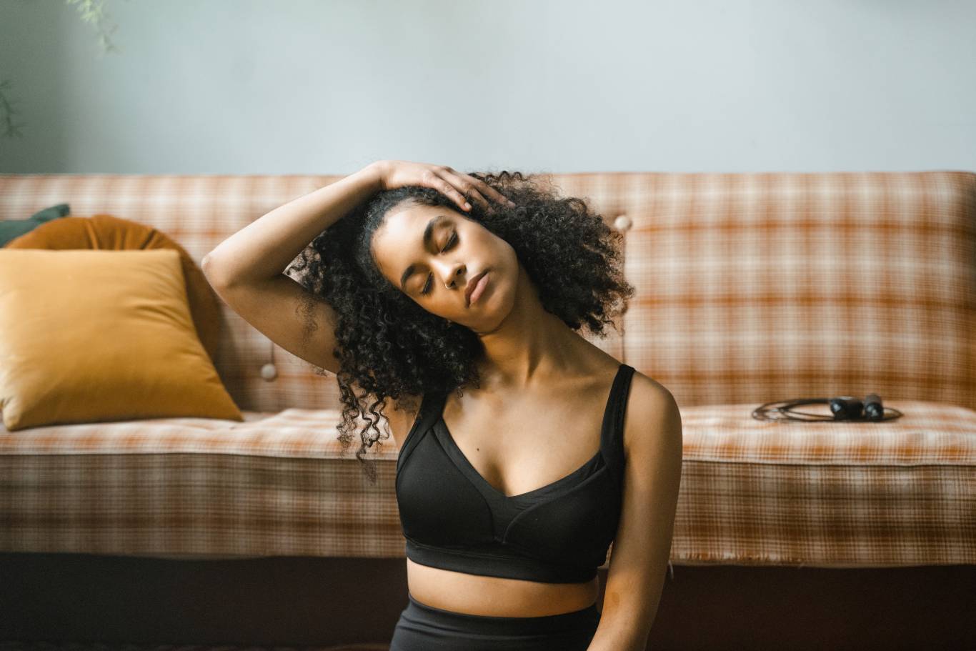 Young woman with curly hair stretches on a plaid sofa, hand resting on her head; warm cushions and headphones create a calm mood.