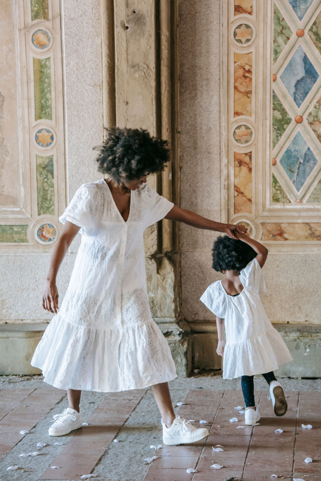 An older child in a white dress guides a young girl in white through a dance pose in an ornate, sunlit hall, conveying warmth and playfulness.