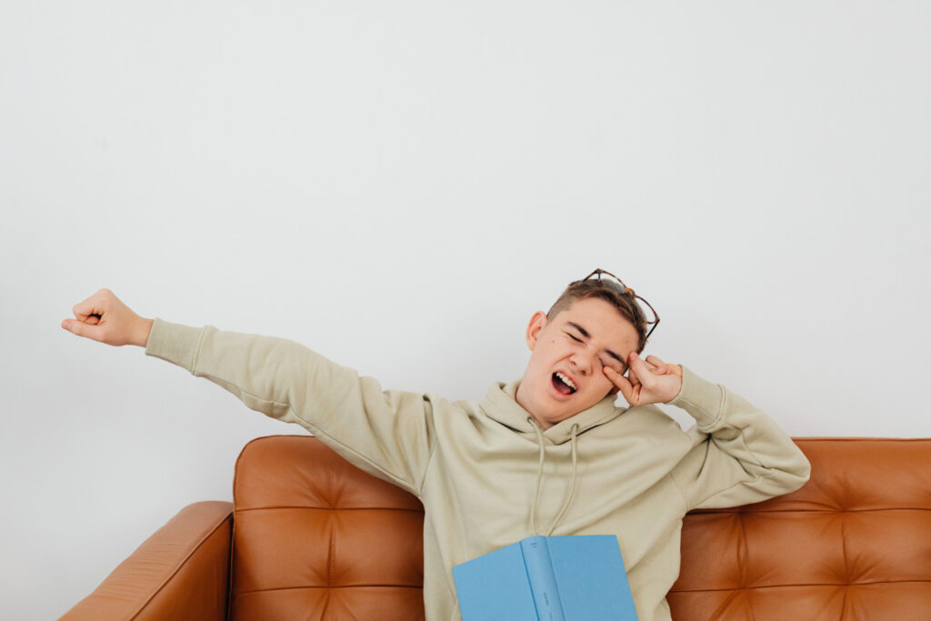 Teenager in a beige hoodie stretches and yawns on a brown leather sofa, eyes closed and glasses askew, with a blue book nearby